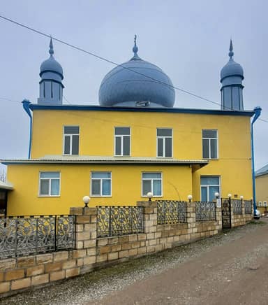 Жёлтое здание мечети с серым куполом и двумя небольшими минаретами в селе Хиль.