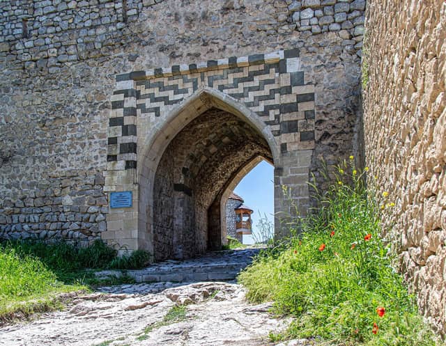 Şuşa qalasının Gəncə qapısı: ağ-qara daşla naxışlanmış sivri tağlı darvaza, yanında lövhə, yolun kənarında qırmızı lalələr