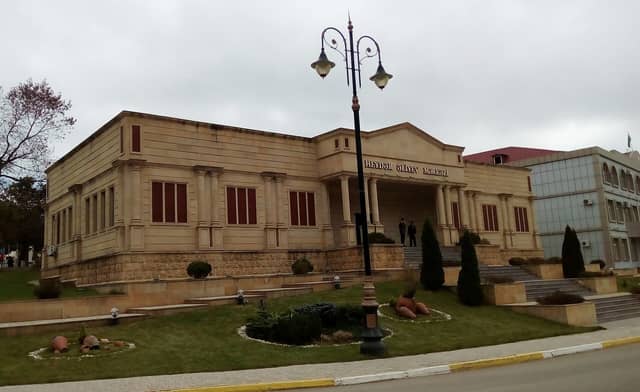 Lerik şəhərində sütunlu girişi olan açıq rəngli daş bina və önündəki yaşıl qazon.