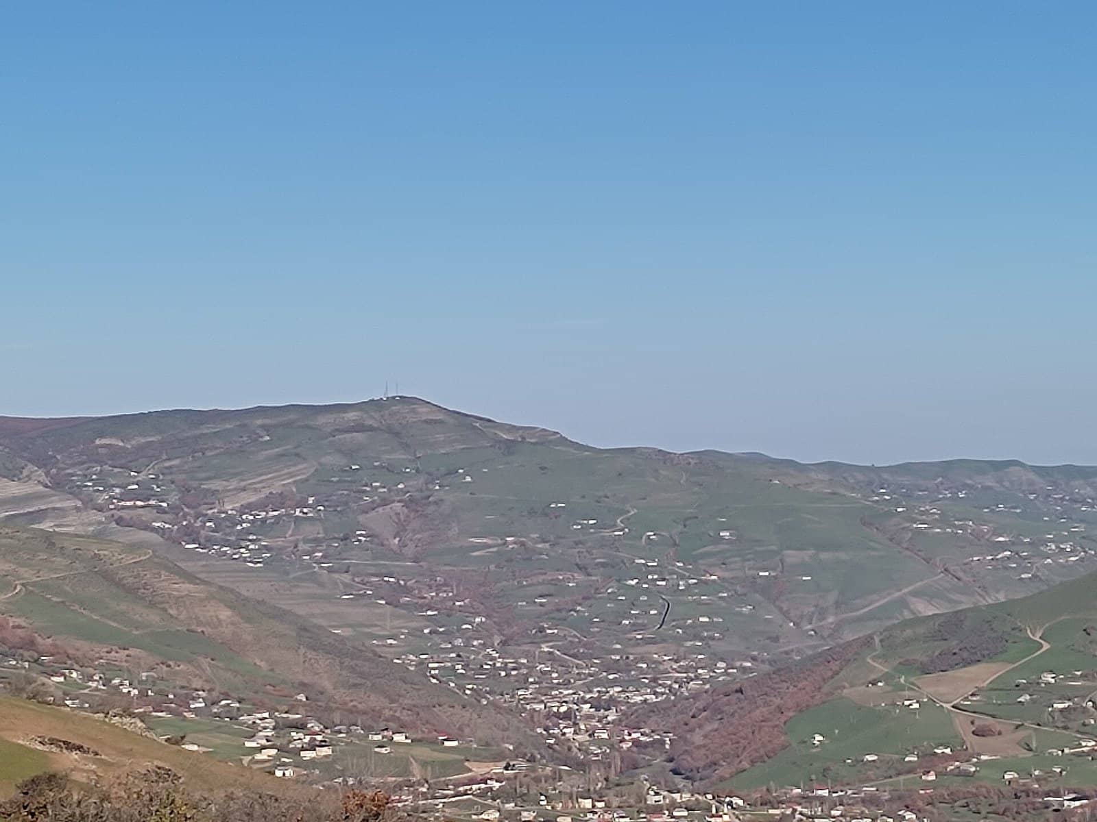 Yardımlı rayonunda dağ yamacları boyunca səpələnmiş kənd evləri və mavi səma altında zirvə.