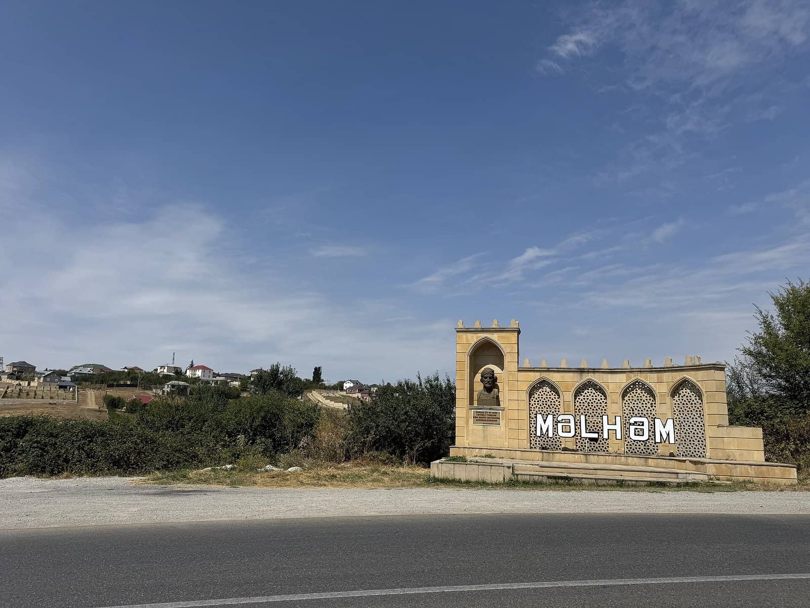 Məlhəm kəndinin girişində adı yazılmış daş abidə və yol, arxada kənd evləri, Şamaxı rayonu