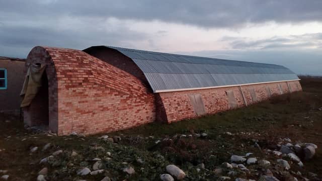 Orta əsr Ağsu şəhəri arxeoloji kompleksi: qazıntı sahəsini örtən kərpic divarlı, tağlı dəmir damlı uzun bina, axşam buludlu səma